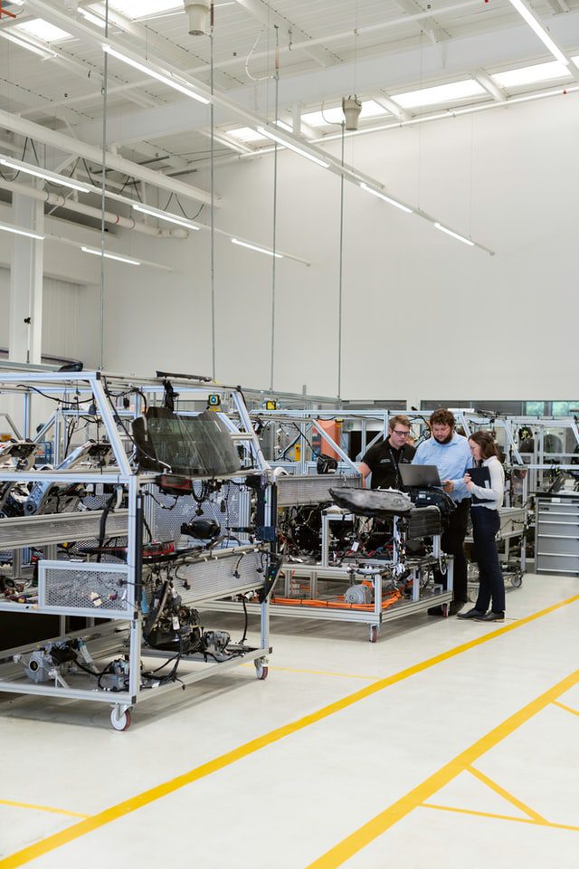 Engineers stand beside vehicle assembly rigs in bright factory, collaborating over laptop while inspecting components.