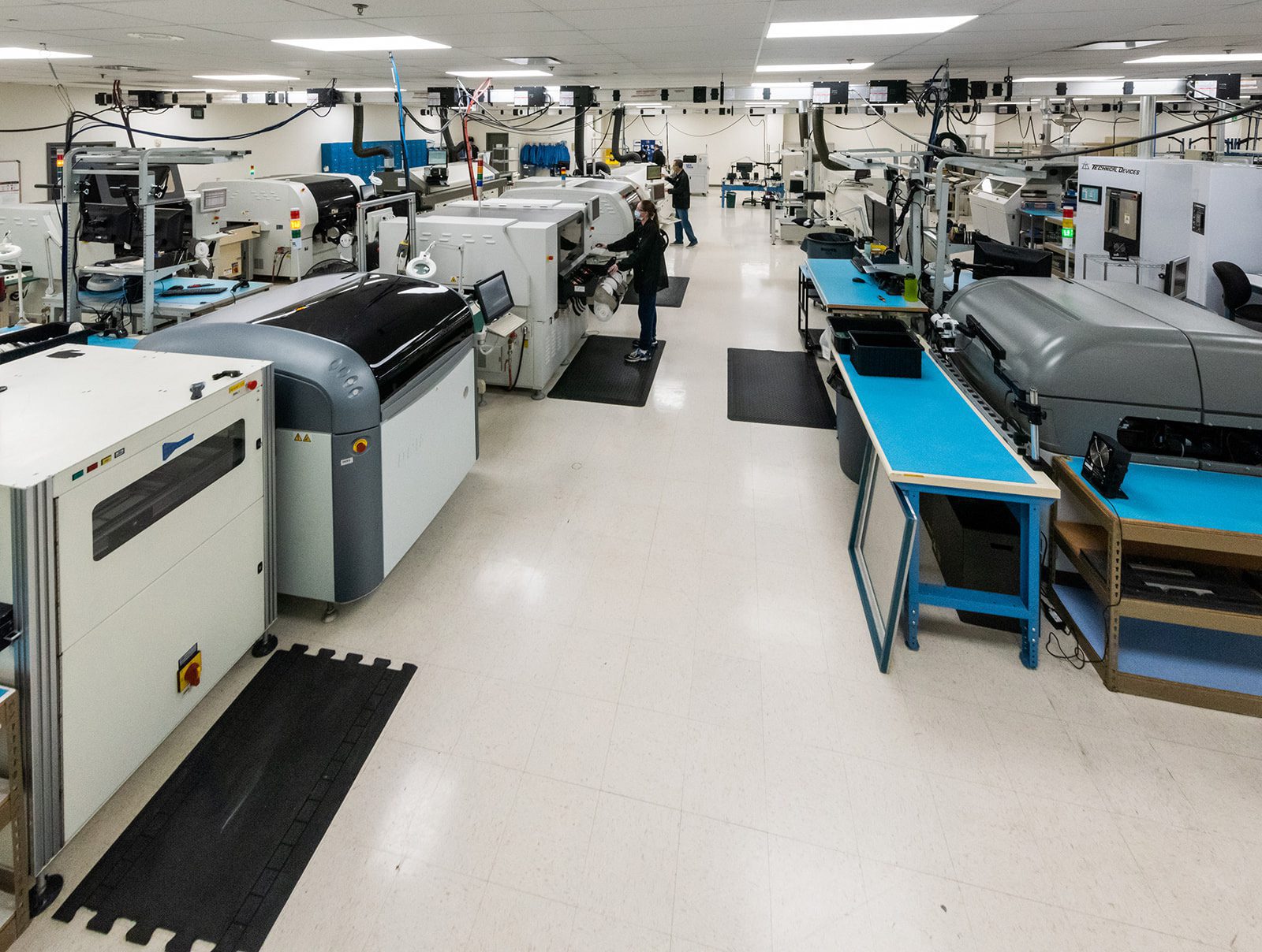 Wide view of electronics manufacturing floor with automated assembly machines, workbenches, and technicians at work.