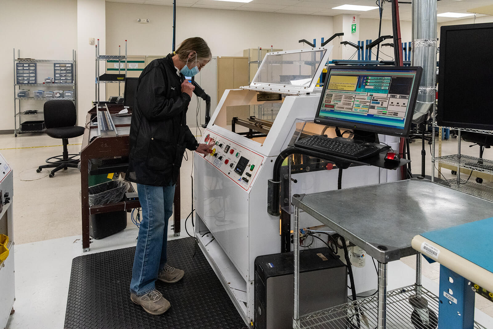 A technician in a lab coat and mask operates complex machinery in a clean, well-organized electronics testing facility.
