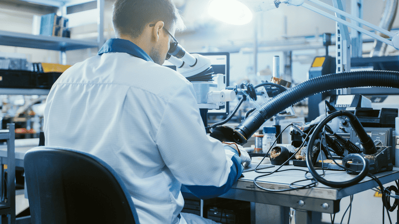 Engineer in a cleanroom using a microscope for precision soldering in an electronics manufacturing facility.