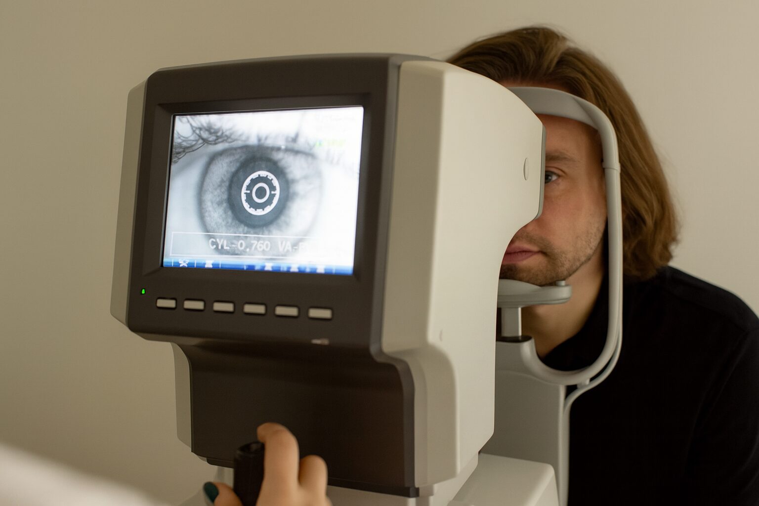 Man undergoing eye exam at optometry clinic, looking into autorefractor machine showing closeup image of eye on screen.