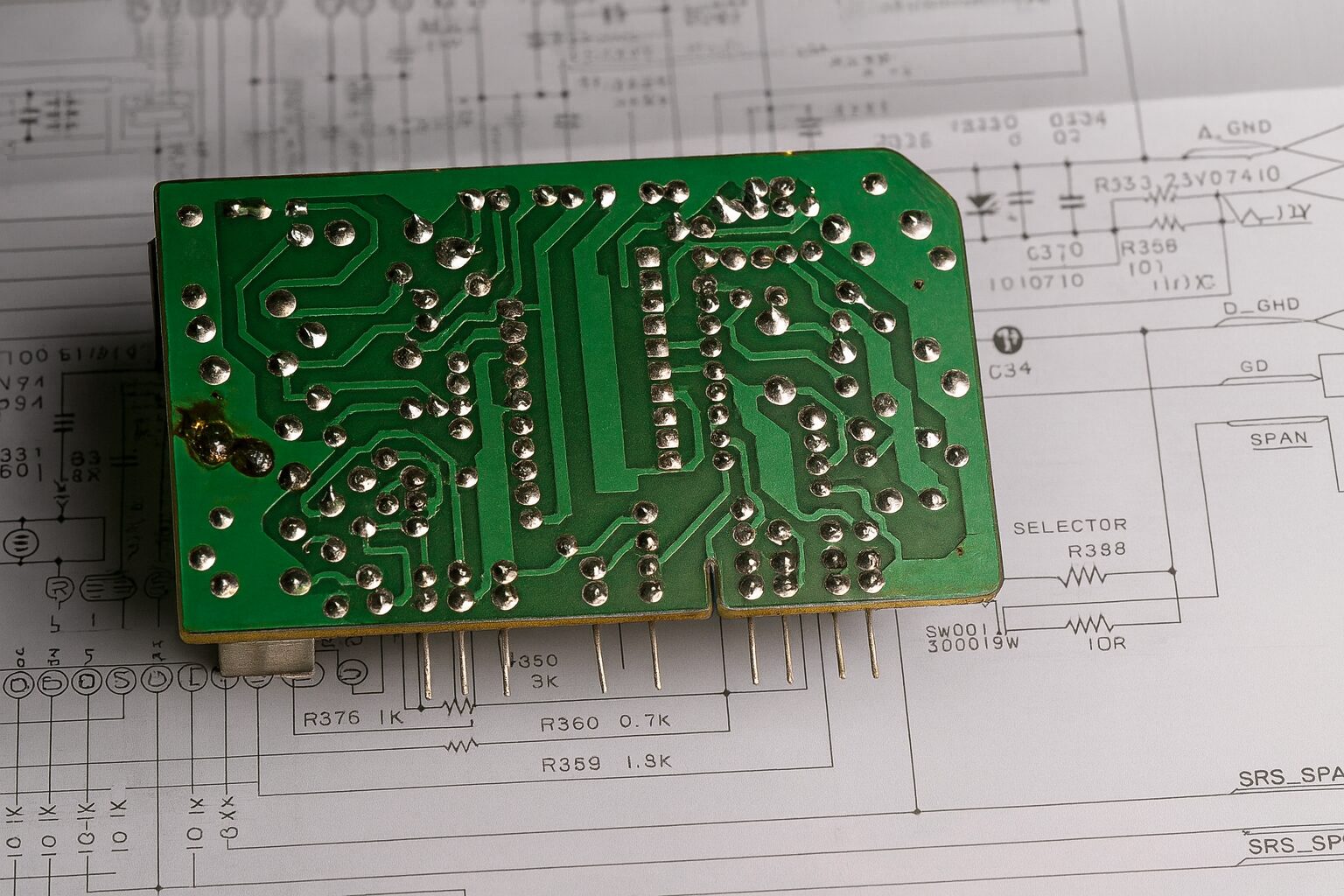 Green printed circuit board with soldered traces lies on top of detailed electronic circuit diagrams and schematics.