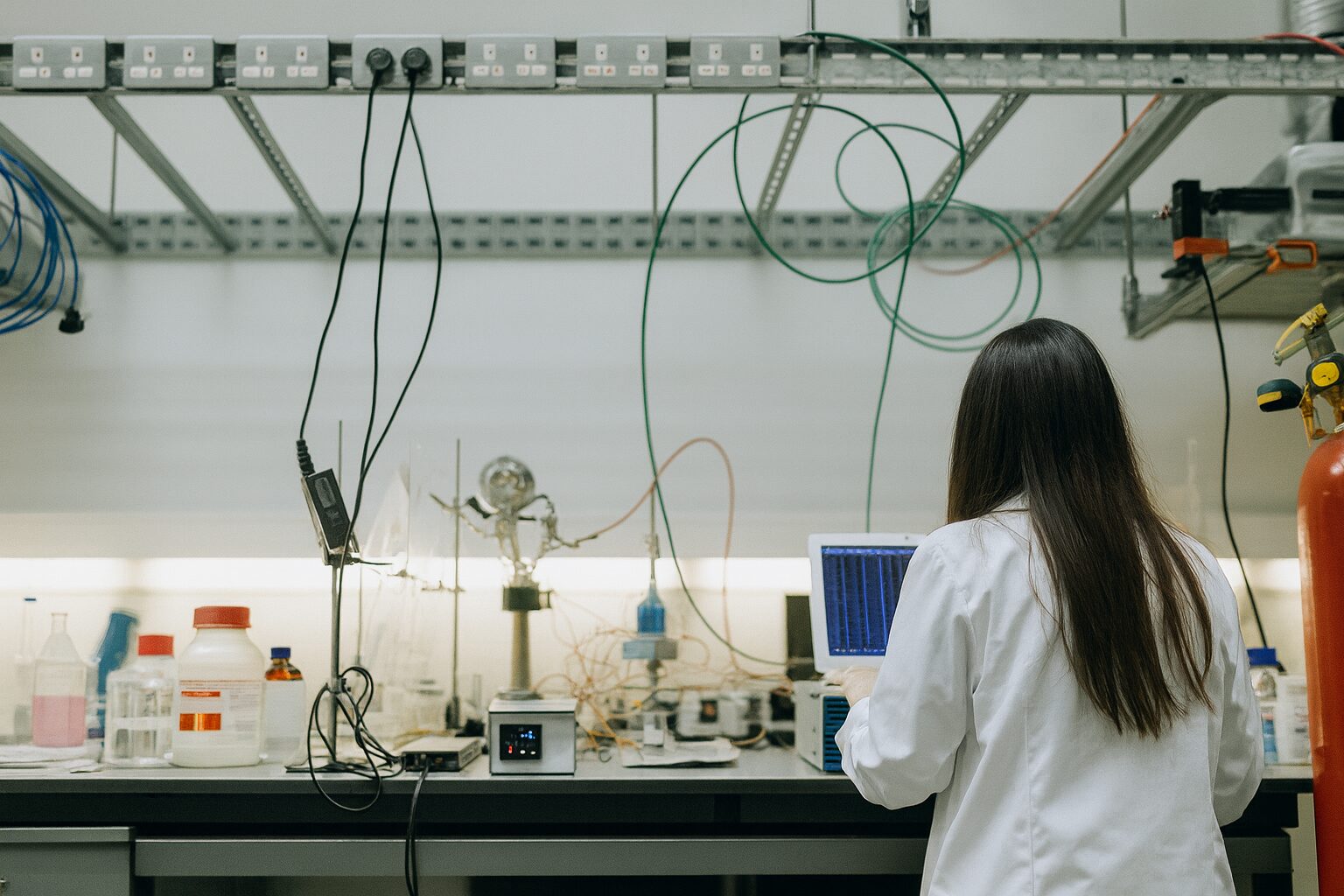Female scientist in lab coat working on laptop at cluttered laboratory bench with chemicals, wires and equipment.