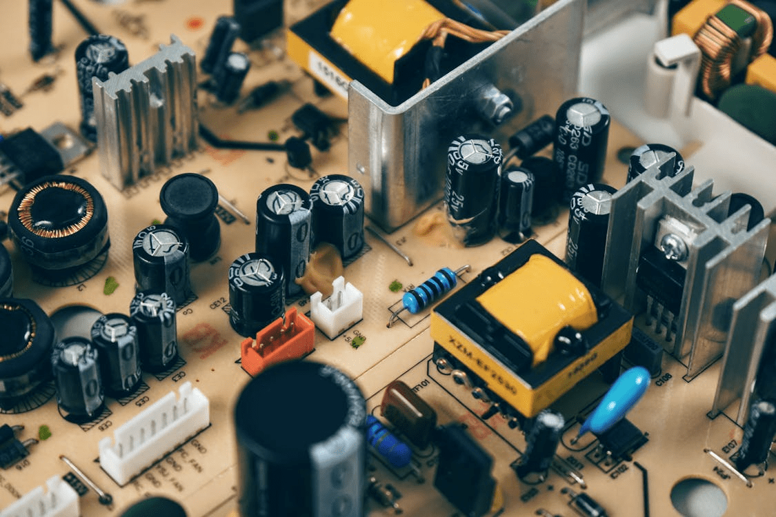 Close-up of electronic circuit board showing capacitors, resistors, heat sinks and components mounted on tan substrate.